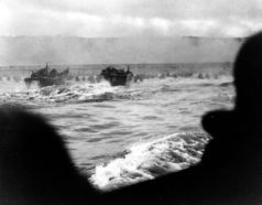 Lanchas de desembarco LCVP desembarcando tropas en la primera oleada de la playa Omaha, a las 6:30 h. La LCVP del extremo izquierdo es del USS Samuel Chase. El fotógrafo, ubicado en el compartimento del conductor de la embarcación, captura las tropas atravesando el mar embravecido y el humo de los bombardeos. En el horizonte, se vislumbran las oscuras alturas detrás de la playa.