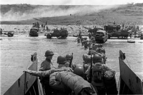 Tropas saliendo de una lancha de desembarco LCVP en Omaha, el 6 de junio de 1944. Obsérvense detrás los vehículos anfibios DUKW entrando en tierra, los semiorugas remolcando cañones anticarro por la orilla, y las filas de hombres que se dirigen tierra adentro.
